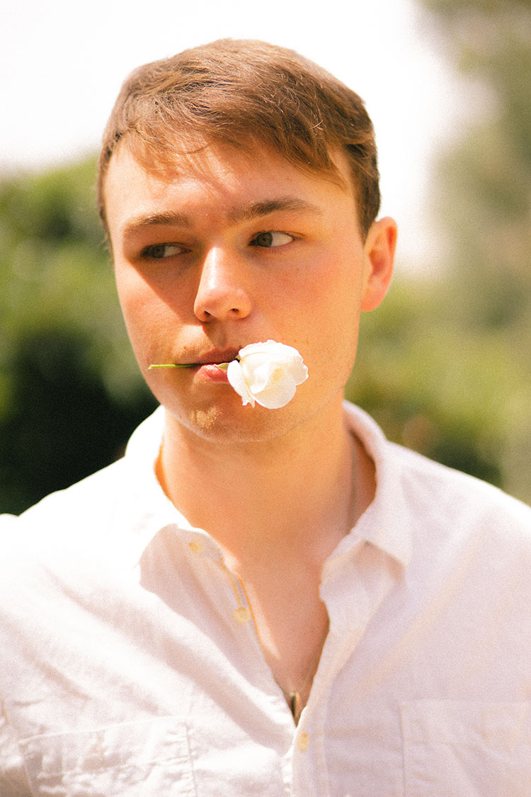 Man in white button-up shirt holds a white flower between his lips